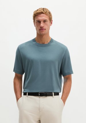 Mann mit kurzen blonden Haaren, der ein blaues kurzärmliges Hemd in beige Hose mit schwarzem Gürtel gesteckt trägt und mit den Händen in den Taschen steht.