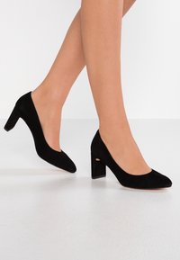 Pumps neri in suede con tacco alto, punta arrotondata e tacco a blocco. La parte superiore ha una texture liscia e un design a scollo basso.