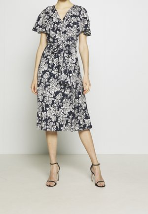 Femme portant une robe noire mi-longue à imprimé floral blanc, manches courtes, col en V, ceinture à la taille et sandales noires à talons hauts avec bride à la cheville.