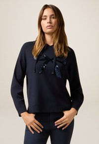 Pull en maille bleu marine avec manches longues, présentant un design de nœud en sequins à l'avant et un ourlet côtelé. Texture lisse, coupe décontractée.