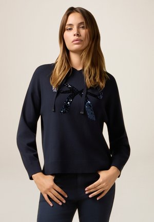 Marineblauer Strickpullover mit langen Ärmeln, der ein Paillettenschleifen-Design auf der Vorderseite und einen gerippten Saum aufweist. Glatte Textur, lockere Passform.