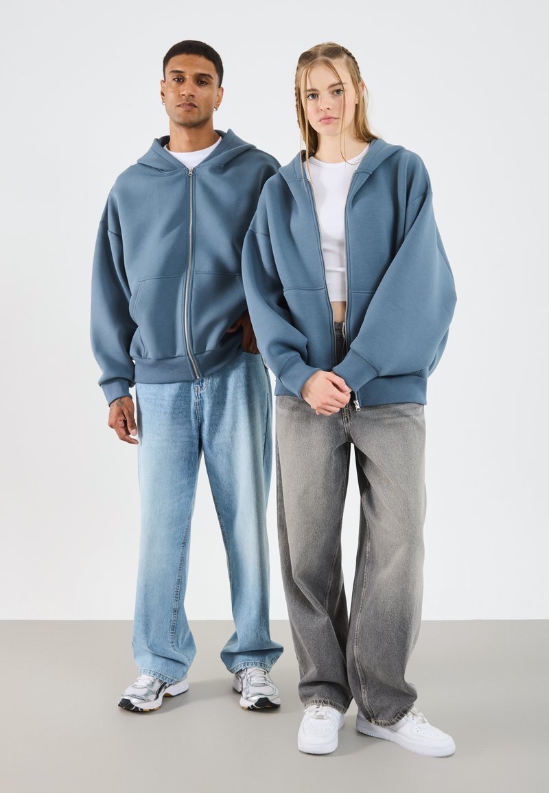 Twee modellen dragen oversized hoodies. De blauwe hoodie heeft een ritssluiting aan de voorkant en grote zakken. Ze combineren deze met licht en donker denim.