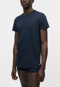 Άνδρας που φοράει μπλε σκούρο κοντομάνικο μπλουζάκι και ταιριαστά σορτς, στέκεται μπροστά σε απλό ανοιχτό γκρι φόντο.