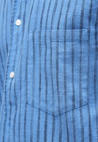 Niebieska koszula z pionowymi czarnymi paskami, białymi guzikami i kieszenią na piersi z dopasowanym wzorem pasków.