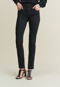Czarne obcisłe jeansy z wysokim stanem i zapięciem na dwa guziki; charakteryzują się gładką fakturą i klasycznym fasonem z pięcioma kieszeniami, zestawione z czarnymi sandałami na obcasie.