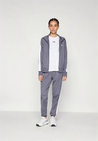 Sivi sportski komplet koji uključuje jaknu na patentni zatvarač s bijelim bočnim detaljima i odgovarajuće hlače. Model nosi bijelu majicu ispod.