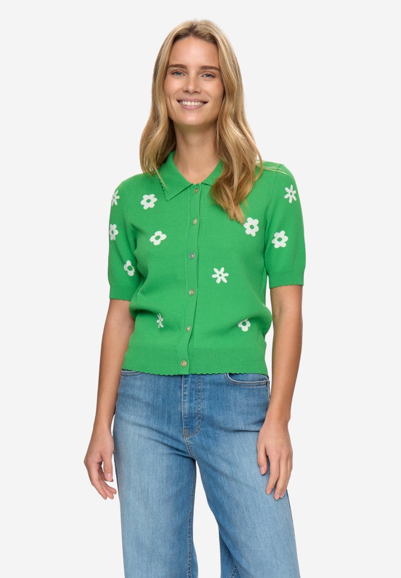 Zelený pletený kardigan s bílými květinovými vzory, krátkými rukávy, zapínáním na knoflíky vpředu a vroubkovaným lemem, kombinovaný s modrými džíny.