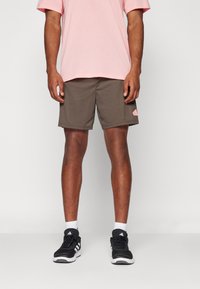 Conjunto masculino que presenta una camiseta de algodón rosa y pantalones cortos deportivos marrones con un parche de logo en el lateral. Combinado con zapatillas deportivas negras y calcetines blancos.