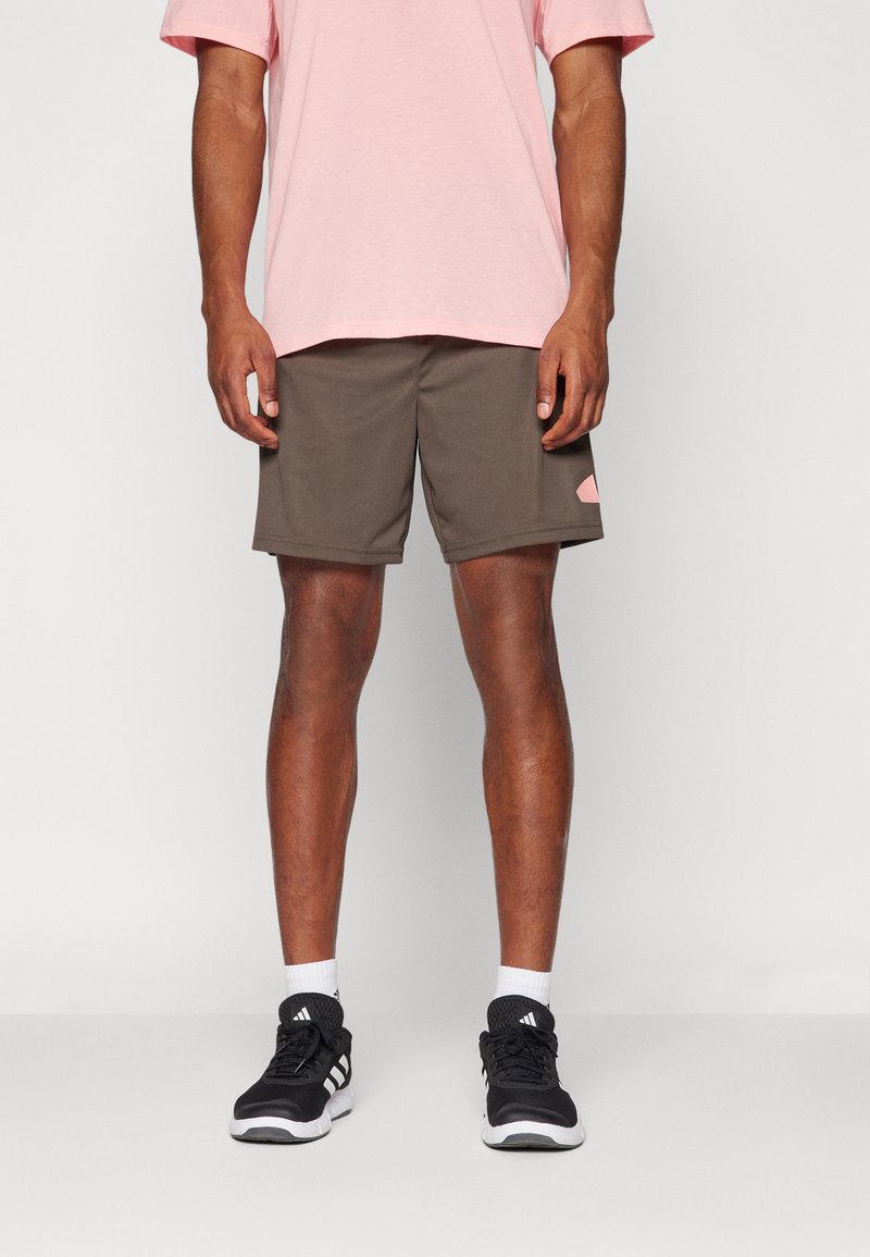 Conjunto masculino que presenta una camiseta de algodón rosa y pantalones cortos deportivos marrones con un parche de logo en el lateral. Combinado con zapatillas deportivas negras y calcetines blancos.