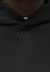 Čierna mikina s kapucňou s klokankovým vreckom a zlatým vyšívaným logom "BOSS" na hrudi. Hladká textúra s kapucňou na šnúrku a rebroványmi manžetami.