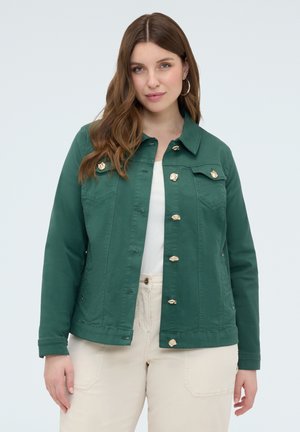 Kobieta o długich brązowych włosach nosi ciemnozieloną zapinaną na guziki kurtkę na białą koszulę i jasnobeżowe spodnie, stojąc na jednolitym tle.