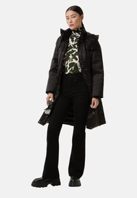Frau trägt einen langen schwarzen Mantel über einem grün-weißen gemusterten Rollkragenpullover, schwarze Hose und klobige schwarze Schuhe, während sie eine Sonnenbrille hält.