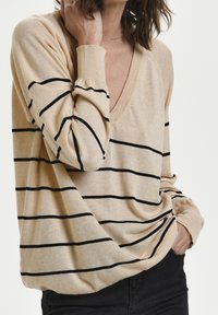 Pull beige à col en V avec rayures horizontales noires, manches longues et coupe décontractée. Texture douce avec poignets et ourlet côtelés.