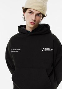 Schwarzer Kapuzenpullover mit Fronttasche, mit weißem Text und Branding auf der Brust. Das Modell trägt eine hellbeige gerippte Mütze.