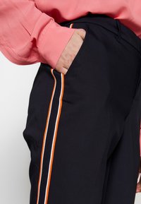 Námořnické kalhoty s bočními pruhy v oranžové, bílé a černé barvě, s přední kapsou a hladkým texturem. Kombinováno s růžovou blůzou.