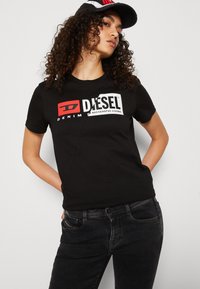 T-shirt en coton noir avec un col rond, arborant un logo Diesel blanc et rouge bien visible sur la poitrine. Porté avec un jean foncé.