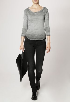 Grijs shirt met lange mouwen, een afgeronde halslijn en drie knopen, gecombineerd met zwarte slim-fit jeans. In één hand wordt een stijlvolle zwarte totebag vastgehouden.