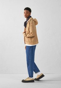 Giubbotto cappuccio marrone chiaro con zip frontale, indossato sopra una maglietta bianca, abbinato a jeans blu a gamba dritta e scarpe beige con lacci.