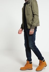 Veste bomber vert olive avec capuche, pull sombre en dessous, jean en denim bleu, et bottes de travail beige avec des accents noirs.