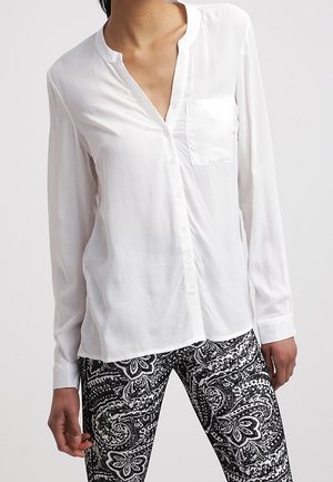 Biała, długorękawowa bluzka zapinana na guziki z kieszenią na piersi, noszona z czarno-białymi spodniami w wzór paisley.