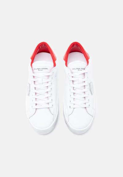 Λευκά αθλητικά παπούτσια με λείο δερμάτινο επάνω μέρος, κόκκινη επένδυση και αντίθετες υφασμάτινες λεπτομέρειες. Περιλαμβάνουν στρογγυλά κορδόνια και λογότυπο.