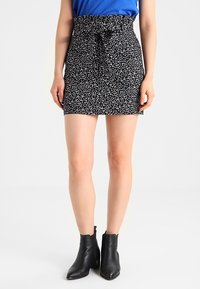 Schwarzer, floraler Minirock mit gebundenem Bund, aus leichtem Material und kleinen weißen Blumendrucken. Kombiniert mit schwarzen Knöchelschuhen.