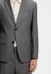 Homme portant une veste de costume gris à fines rayures et un pantalon assorti sur une chemise blanche à col, debout devant un fond uni.