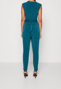 Mujer de pie de espaldas a la cámara, vistiendo un mono sin mangas en color azul verdoso atado en la cintura y sandalias de tacón negro.