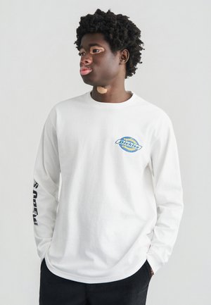 Tricou alb cu mâneci lungi, cu un logo albastru circular pe piept și text negru îndrăzneț pe mâneca stângă; realizat din țesătură de bumbac.