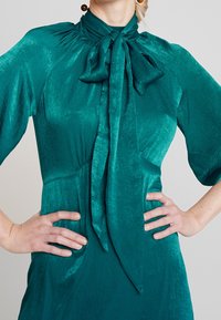 Teal satin klänning med hög krage och rosett, korta ärmar och figursydd liv. Slät textur med subtil lyster.