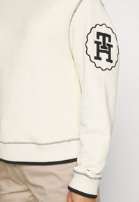 Maglione crema con tessuto testurizzato, dettagli di cucitura nera, polsini e bordo a coste, e un logo circolare nero sulla manica sinistra.