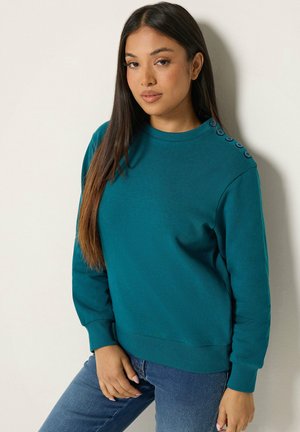 Junge Frau mit langen dunklen Haaren, die einen tealfarbenen Pullover mit Knöpfen an der Schulter und blaue Jeans trägt, steht vor einer hellen Wand.