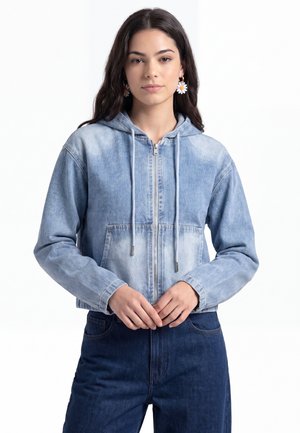 Jeune femme aux longs cheveux foncés portant des boucles d'oreilles marguerite, un sweat à capuche zippé en jean bleu clair et un jean bleu foncé, debout, les mains jointes.