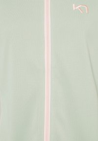 Jasnozielona sportowa kurtka o gładkiej fakturze, z centralnym różowym zamkiem błyskawicznym i małym haftowanym logo w górnym rogu.