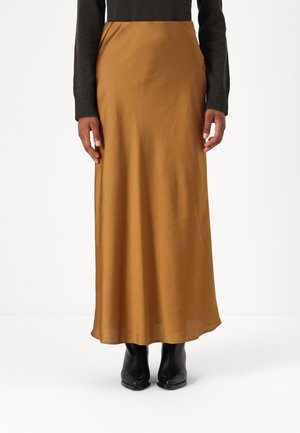 Person iført et langt sennepsfarvet skørt, sorte støvler og en mørk langærmet top, stående oprejst på en hvid baggrund.