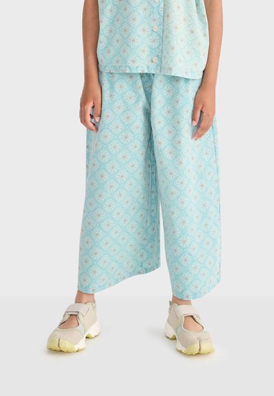 Barn iført lyseblå, mønstrede bukser med brede ben og matchende top, stående med beige sneakers på en ensfarvet lys baggrund.