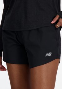 Pantalones cortos deportivos negros con una textura suave. Presenta un logo plateado de New Balance en la parte inferior derecha. Diseñados para la comodidad y el rendimiento.