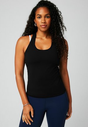 Woman with long curly hair wearing a black tank top, white sports bra strap, blue leggings, silver earrings, bracelet, and rings, standing against gray background.