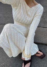 Robe tricotée blanche avec un motif en losanges, manches longues et décolleté ouvert. Portée avec des sandales noires et un gros bracelet clair au poignet.