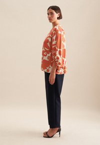 Maglia a maniche lunghe colorata con un motivo floreale, caratterizzata da maniche arricciate; abbinata a pantaloni scuri e sandali con tacco neri.