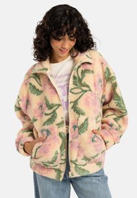 Fleece zip-up jakke med blomsterdesign i rosa og grønn på en kremfarget bakgrunn. Den har krage og to frontlommer.