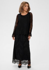 Czarna koronkowa sukienka z długimi przezroczystymi rękawami, zdobiona skomplikowanymi wzorami kwiatowymi i falbanistym dołem, noszona na jednolitej czarnej halka.