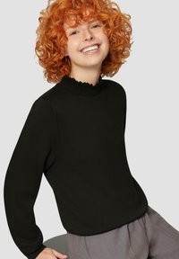 Zwarte trui met een hoge, gerimpelde halslijn. Zachte textuur, lange mouwen, gecombineerd met grijze broeken. Model heeft krullend oranje haar en glimlacht.