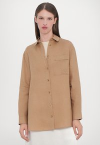 Tan button-up shirt with a collar, long sleeves, a patch pocket, and subtle texture. Features brass buttons and embroidered initials.