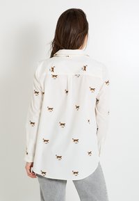 Chemise blanche à boutons avec broderie de petits chats marron clair sur tout le vêtement, dotée de manches longues et d'un ourlet incurvé. Empiècement au dos avec détail de pli.