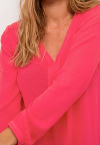 Blusa lunga a maniche lunghe di un rosa brillante, con scollatura a V e tessuto texturizzato. Presenta una vestibilità comoda con maniche leggermente arricciate ai polsi.