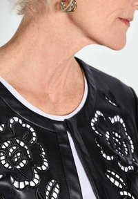 Zwarte leren jas met bloemmotieven in reliëfontwerp en witte accenten, een ronde halslijn en een gladde textuur. Witte shirt zichtbaar eronder.