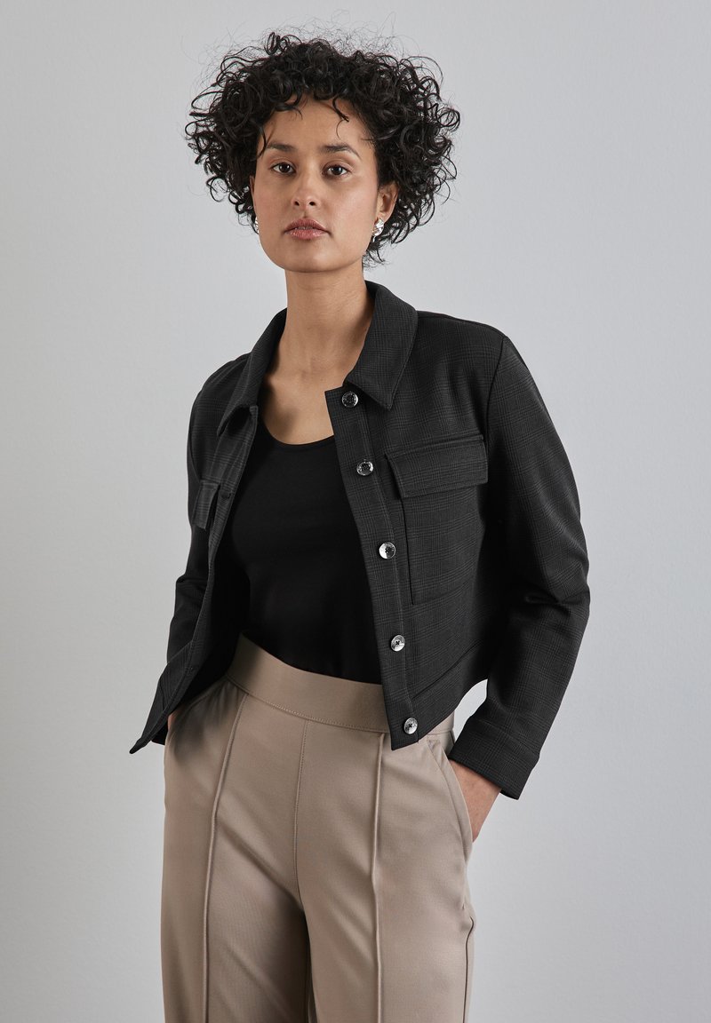 Zwarte cropped jasje met een textuurpatroon, zilveren knopen en een borstzak, gecombineerd met beige hoge taillebroek en een zwart top.