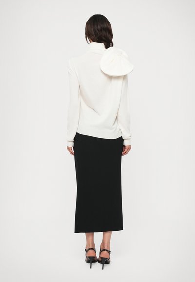 Balts džemperis ar augstu apkakli un unikālu savērtu detaļu aizmugurē, kombinēts ar melnu ceļgalu garuma svārku un melniem augstpapēžu sandaliem.