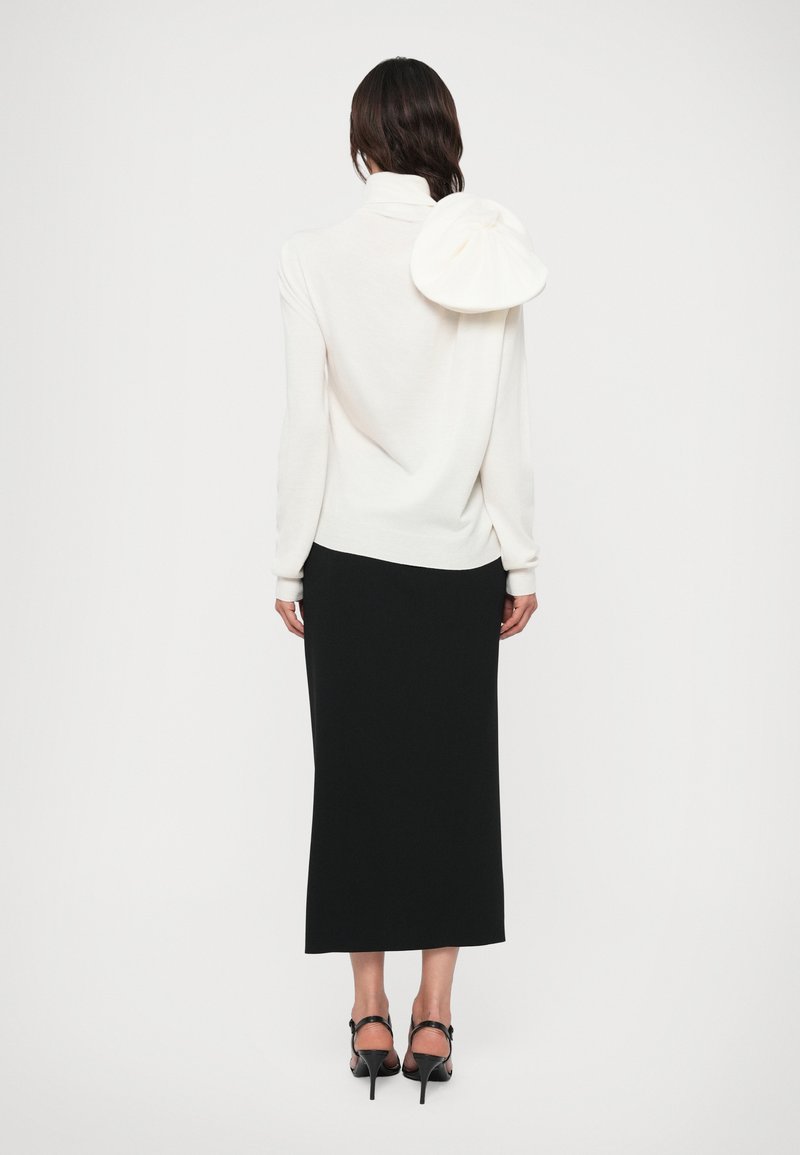 Maglione bianco con collo alto e un particolare dettaglio arricciato sulla schiena, abbinato a una gonna nera lunga fino alla caviglia e sandali neri con tacco.